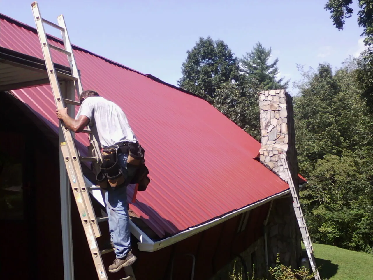 Roofer on a ladder beside a red metal roof and stone chimney — Soffit & Fascia Repair by Roofing Brothers in Grantham